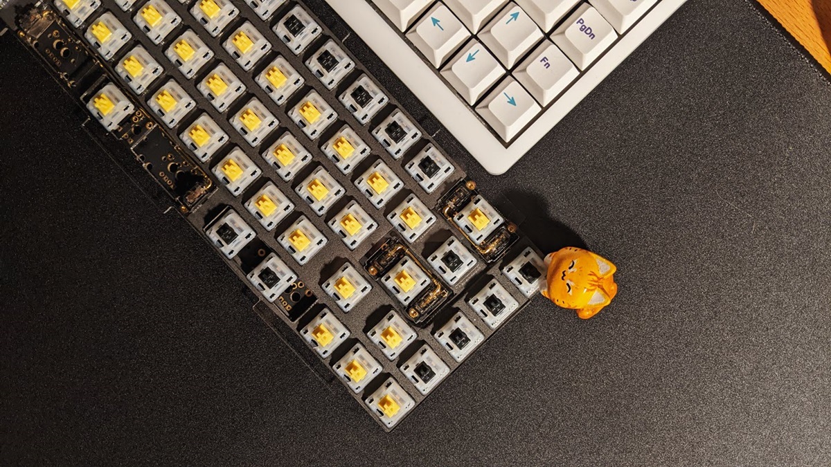A top-down view of a partially disassembled computer keyboard with exposed yellow switches sitting on a black desk mat, with a small orange cat figurine posed at its corner.