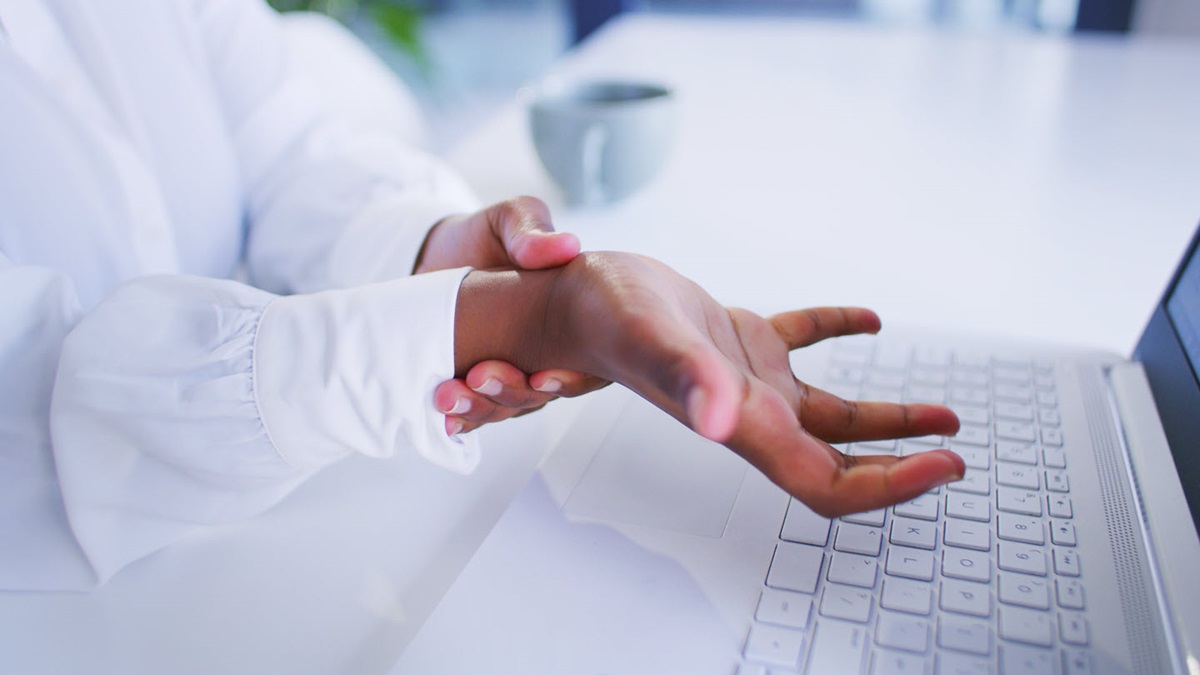 Close-up of a person holding their wrist while seated at a laptop.