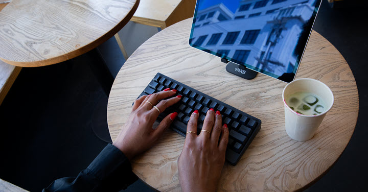 Woman typing on a wireless black keyboard on a coffeehouse table with a beverage and a screen. 