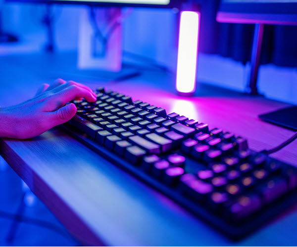 Close-up of a keyboard on a desk lit in purple and violet hues. A person's hand sits perched over the left side of the board.